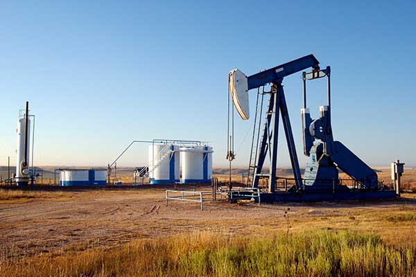 An oil pump jack and white storage tanks on a flat open plain under a clear blue sky.